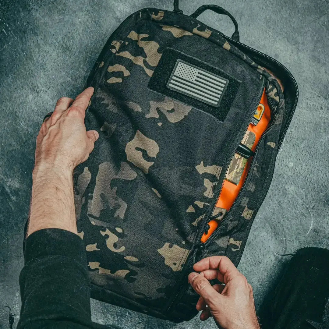 Black camouflage backpack with American flag patch being opened by hands.