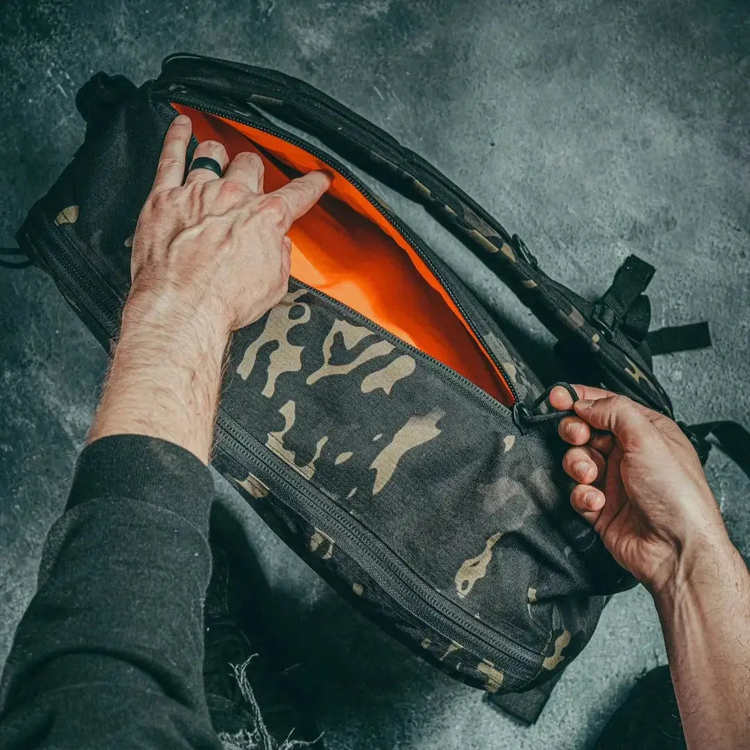 Hands opening a black camouflage backpack with an orange interior.