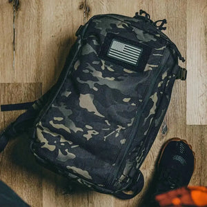Camouflage backpack with American flag patch and shoe on wooden floor.