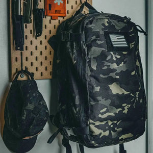 Camouflage backpack hanging on a pegboard with knives and a cap.