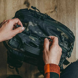 Hands opening zippers on a camouflage 24 Hour Plus backpack.