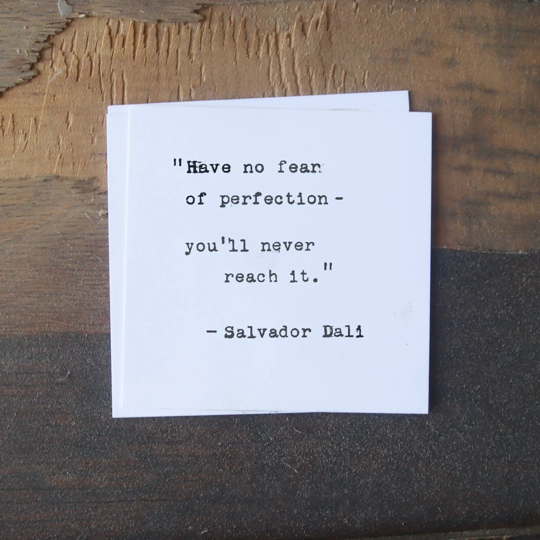 A white card with a Salvador Dali quote sits on a textured wooden surface.