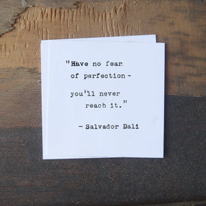 A white card with a Salvador Dali quote sits on a textured wooden surface.