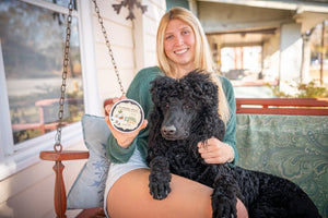 Woman holding almond milk arnica salve with a black poodle on a porch swing.