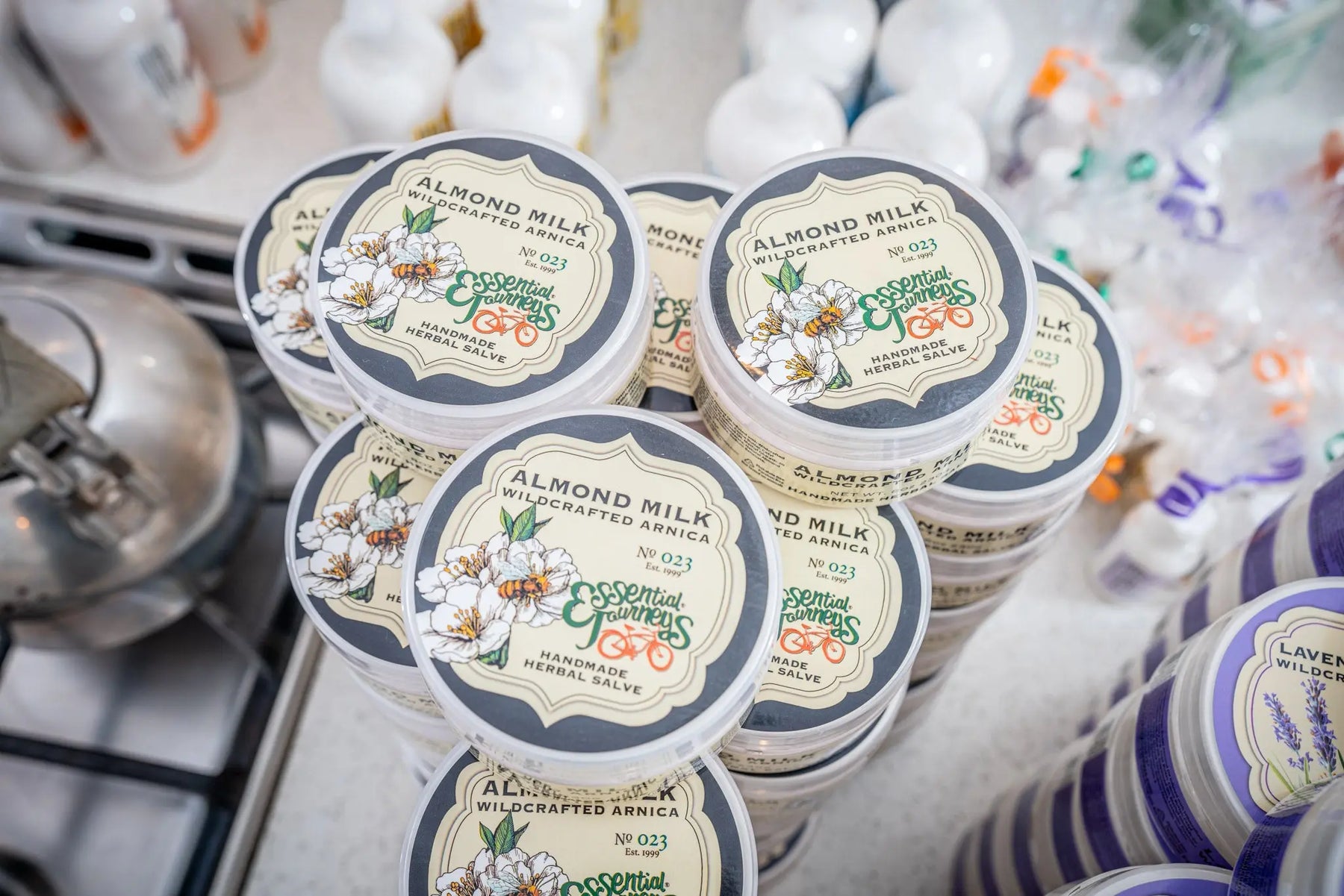 Stack of Almond Milk Wildcrafted Arnica herbal salve containers with visible Essential Journeys branding and floral labels.