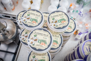 Stack of Almond Milk Wildcrafted Arnica herbal salve containers with visible Essential Journeys branding and floral labels.