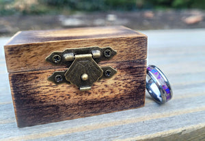Wooden keepsake box with a metal cremation ash ring featuring amethyst opal inlay resting against it on a wooden surface.