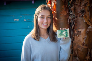 Young woman holding a rectangular bar of green and white swirled soap near a tree.