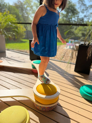 A child in a blue dress stands on a yellow balance board with a multi-colored speckled base on a wooden deck.