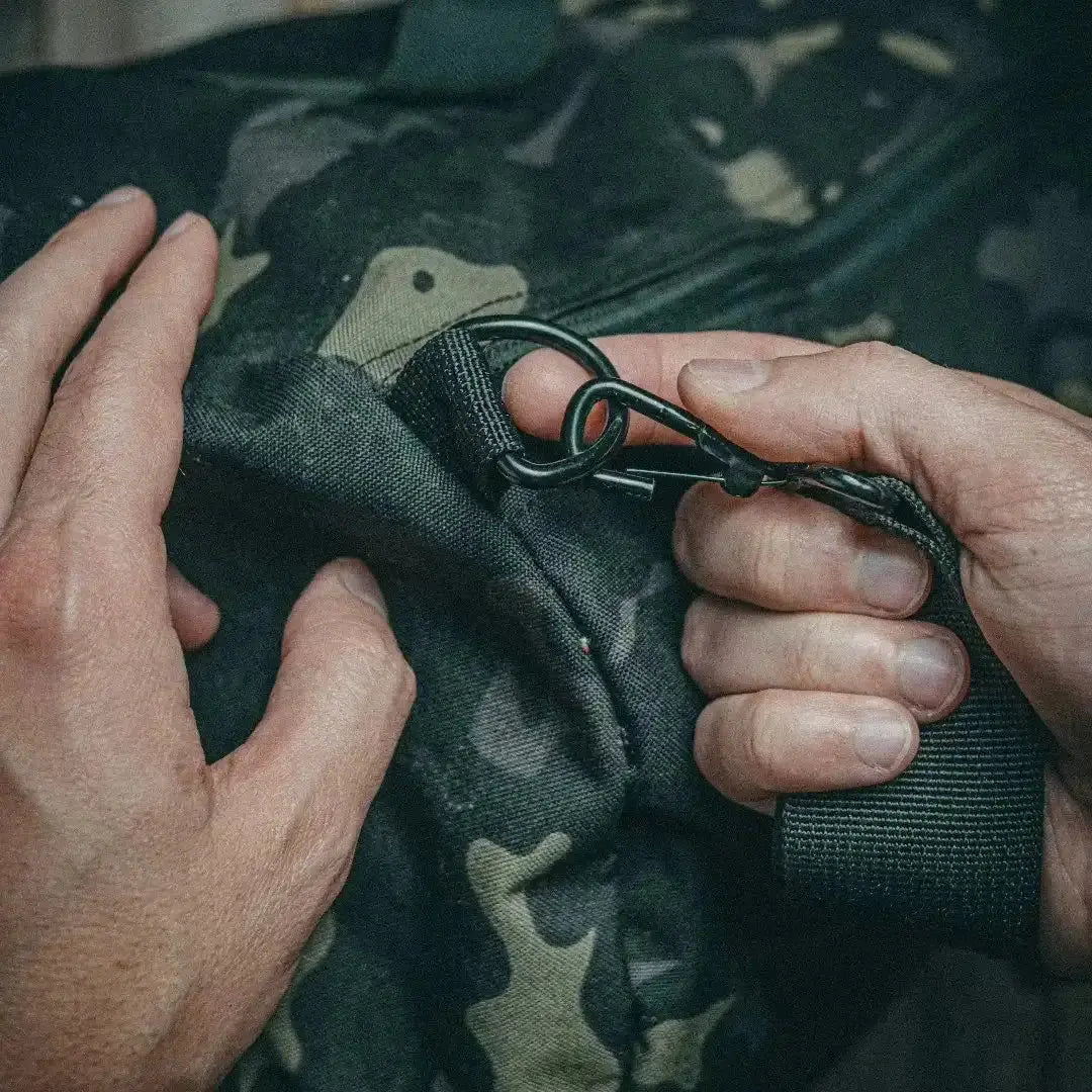 Hands attach a black carabiner clip to a camouflage duffle bag strap.