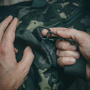 Hands attach a black carabiner clip to a camouflage duffle bag strap.