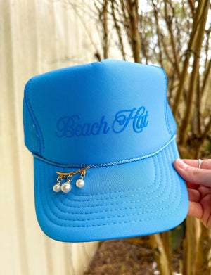 Columbia blue trucker hat with "Beach Hut" cursive text and pearl dangle accessory on brim held by a hand.