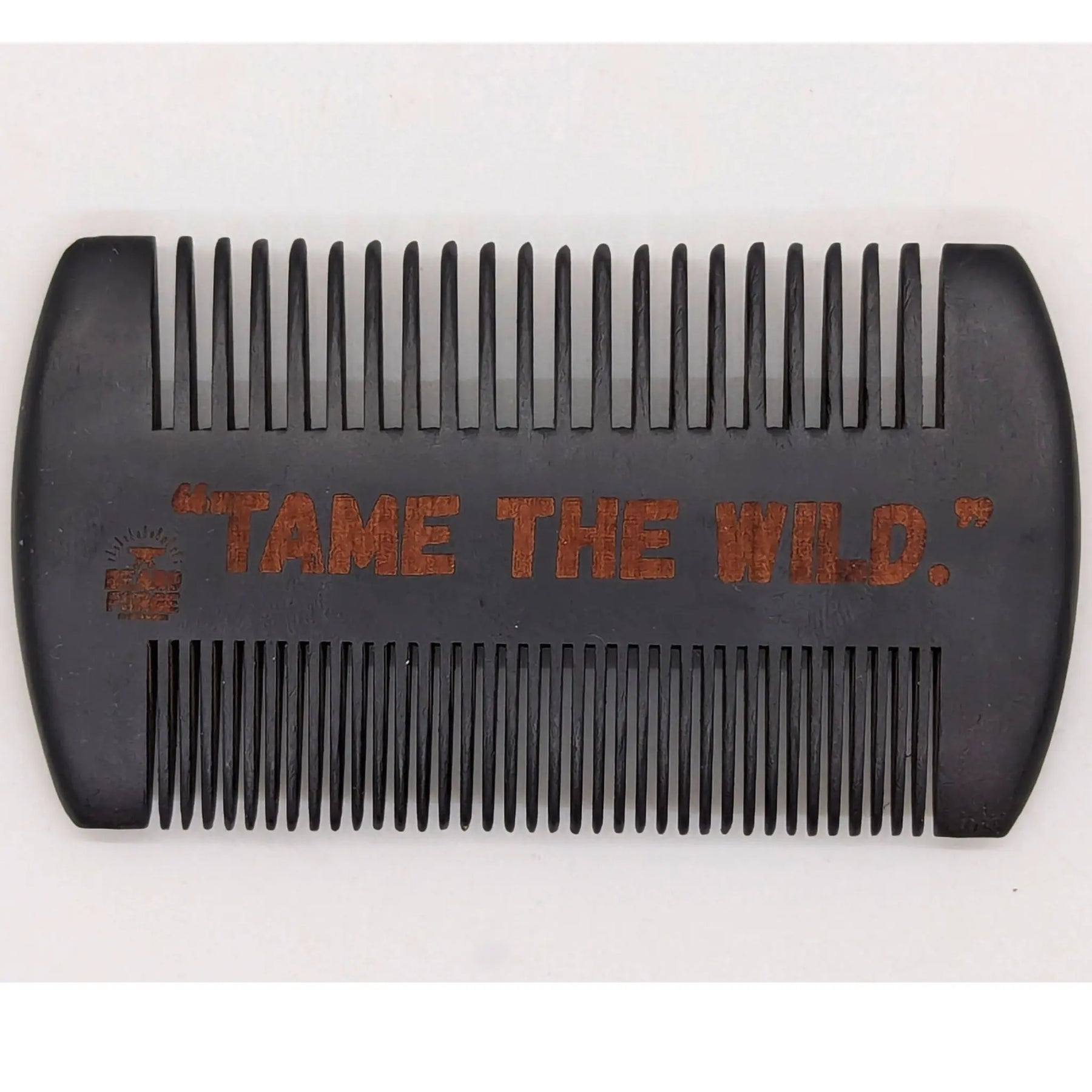 Wooden beard comb with \"Tame The Wild\" engraved on it, showing comb's teeth and overall shape. A grooming tool for beard care.