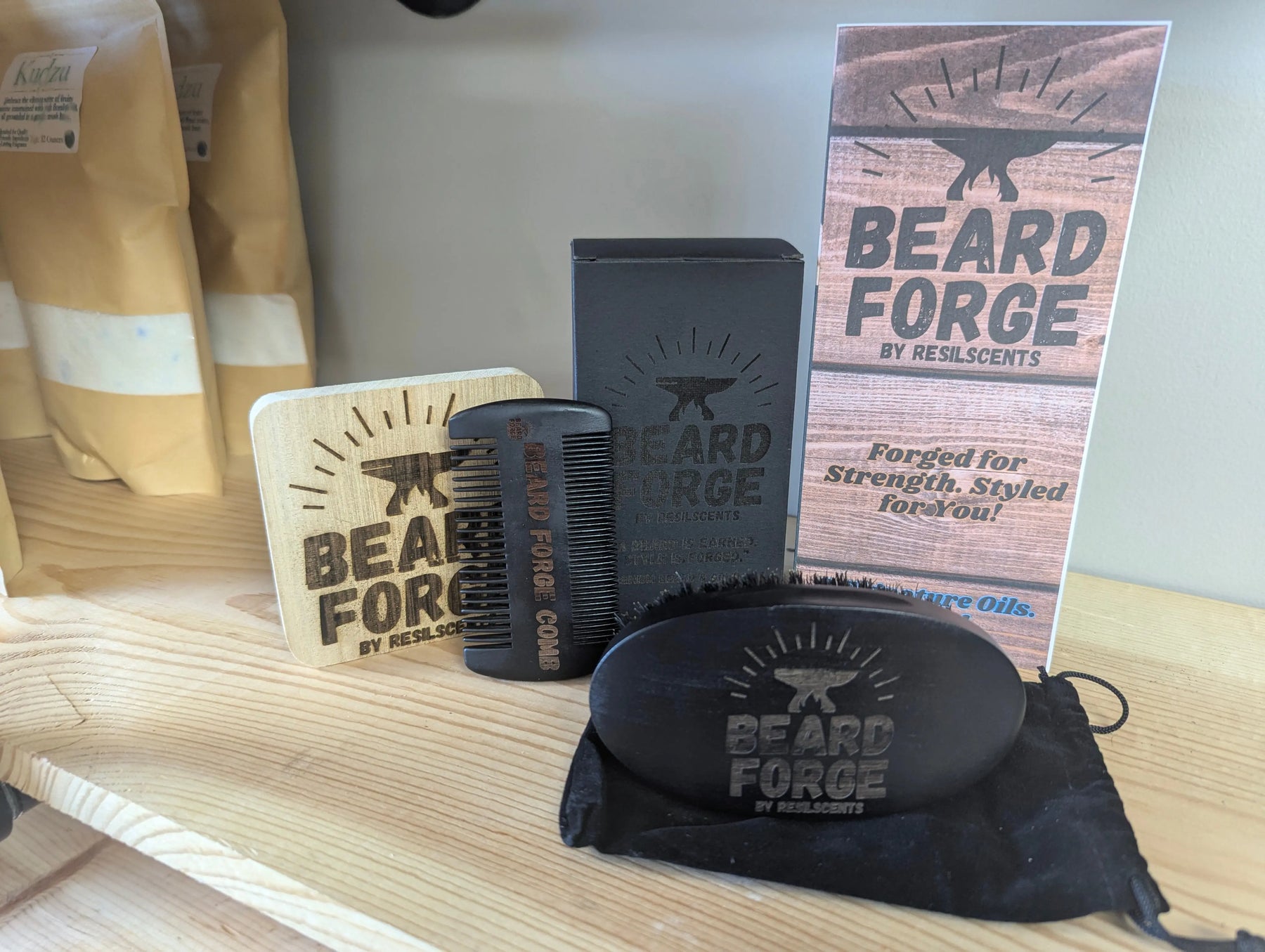 Beard Forge grooming set on a wooden shelf, featuring a brush, comb, box, pouch, and promotional stand with the logo.