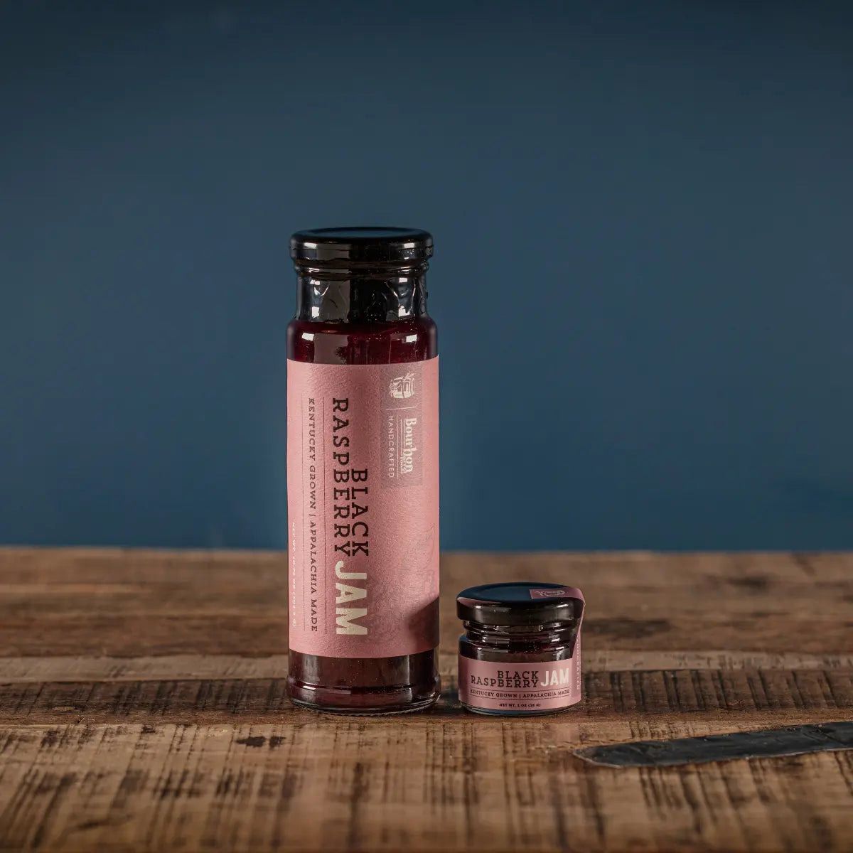 Two glass jars of black raspberry jam with bourbon label on wooden table.