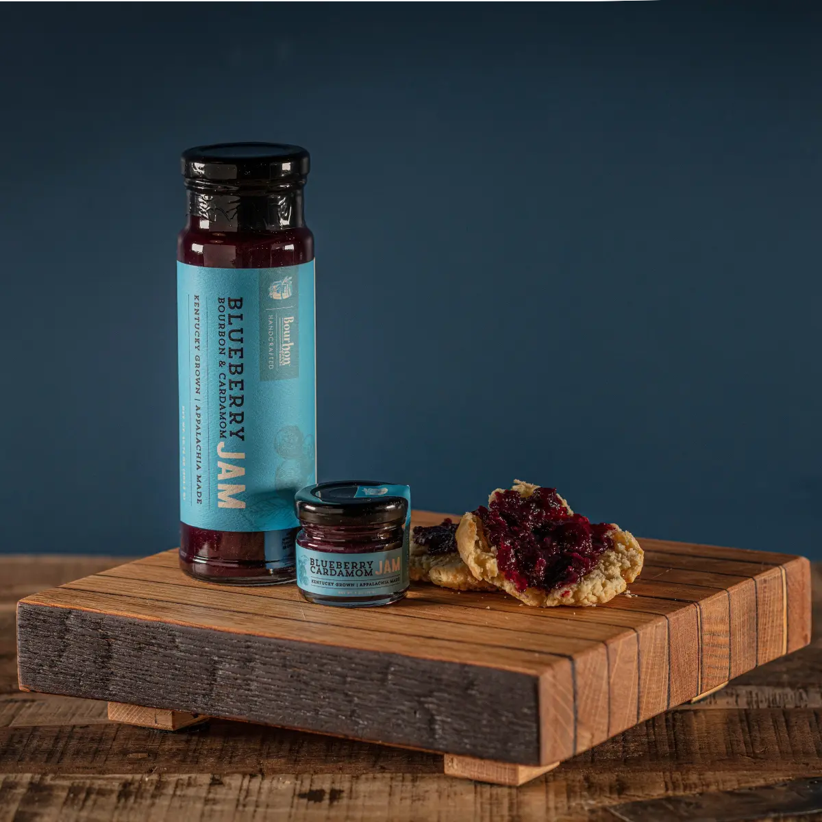 Two glass jars of blueberry jam, one open with jam on bread, on a wooden board.