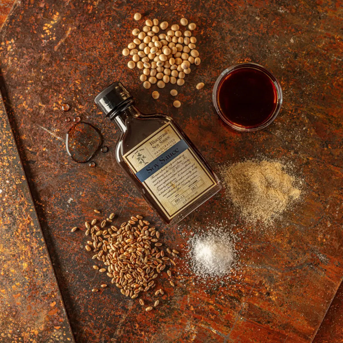 Bottle of Bluegrass Soy Sauce with raw soybeans, wheat, and salt on a textured surface.