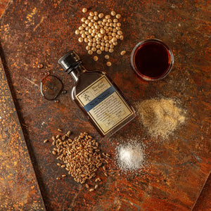 Bottle of Bluegrass Soy Sauce with raw soybeans, wheat, and salt on a textured surface.
