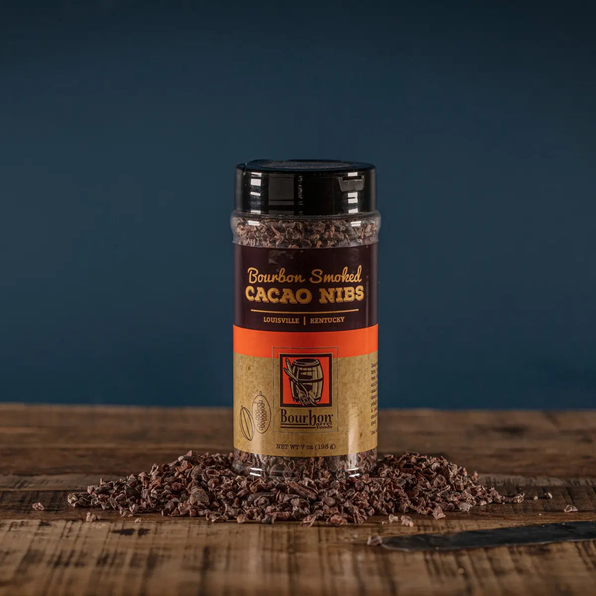 Bourbon Smoked Cacao Nibs in a clear container with scattered cacao nibs on a wooden surface.