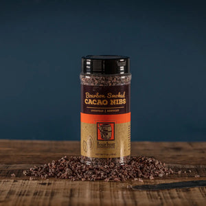 Bourbon Smoked Cacao Nibs in a clear container with scattered cacao nibs on a wooden surface.
