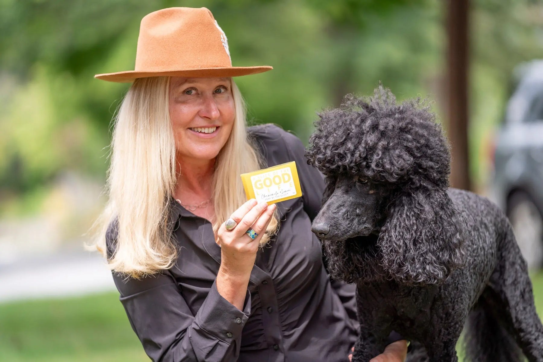 Woman in hat holds chamomile lemon soap next to black poodle