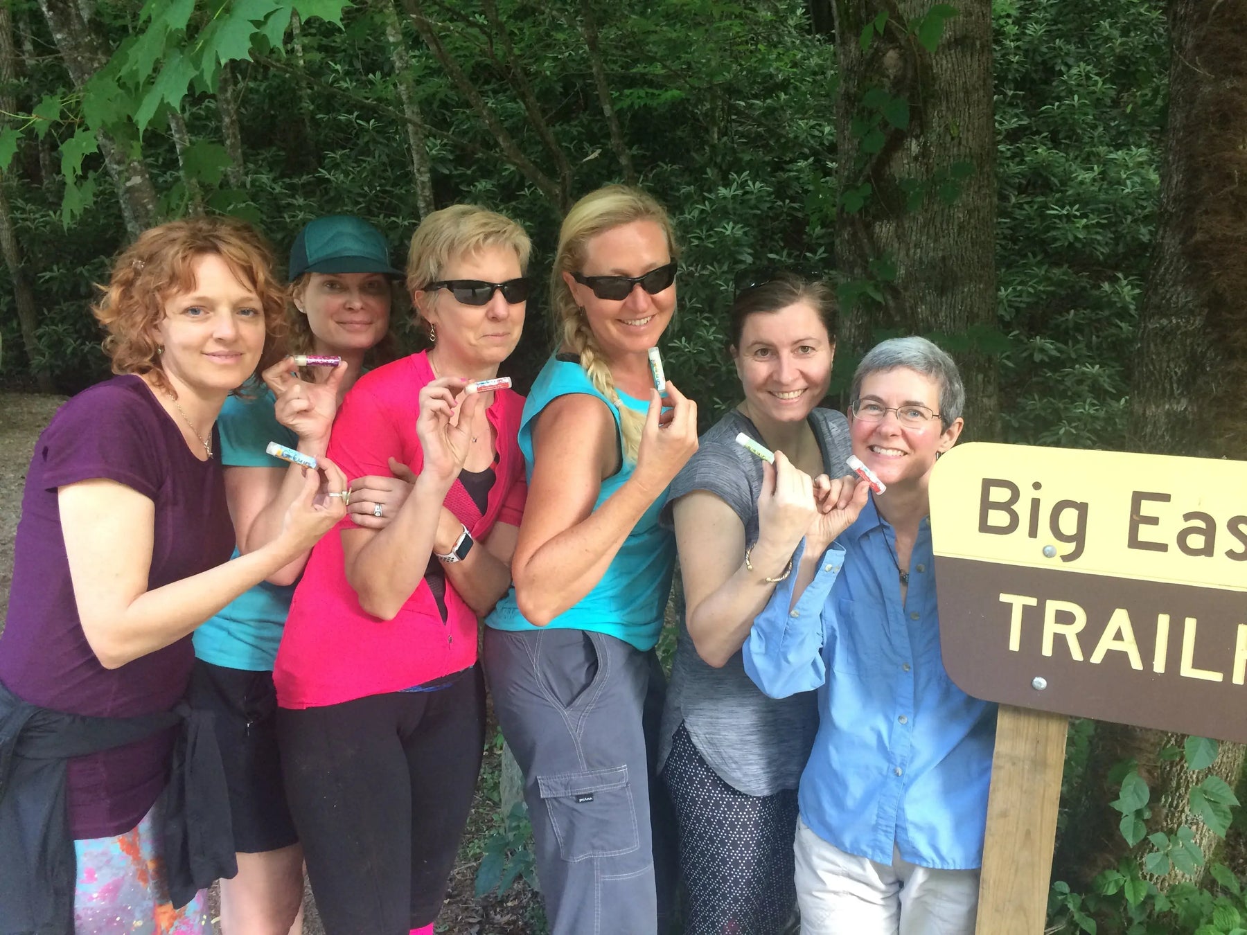 Group of women holding lip balm tubes near Big East Trail sign in forest.