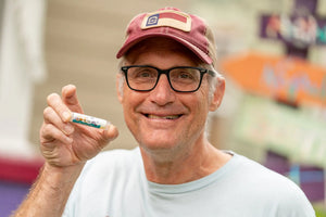 Man with glasses and hat holding "COASTING COCONUT SPF 15 LUBE FOR THE LIPS" lip balm.