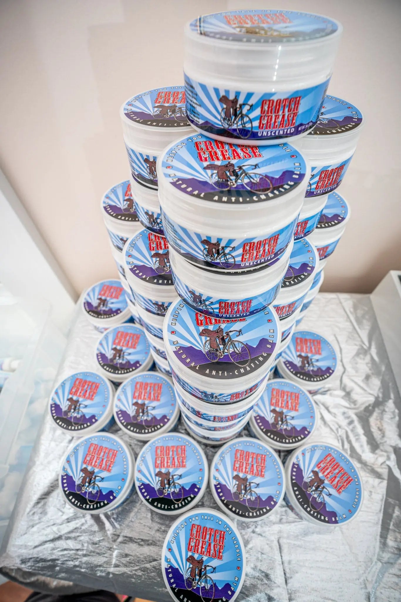 Stack of Crotch Grease unscented anti-chafing balm tubs with cyclist graphics.