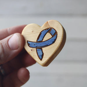 Hand holding a wooden heart with a blue ribbon graphic.