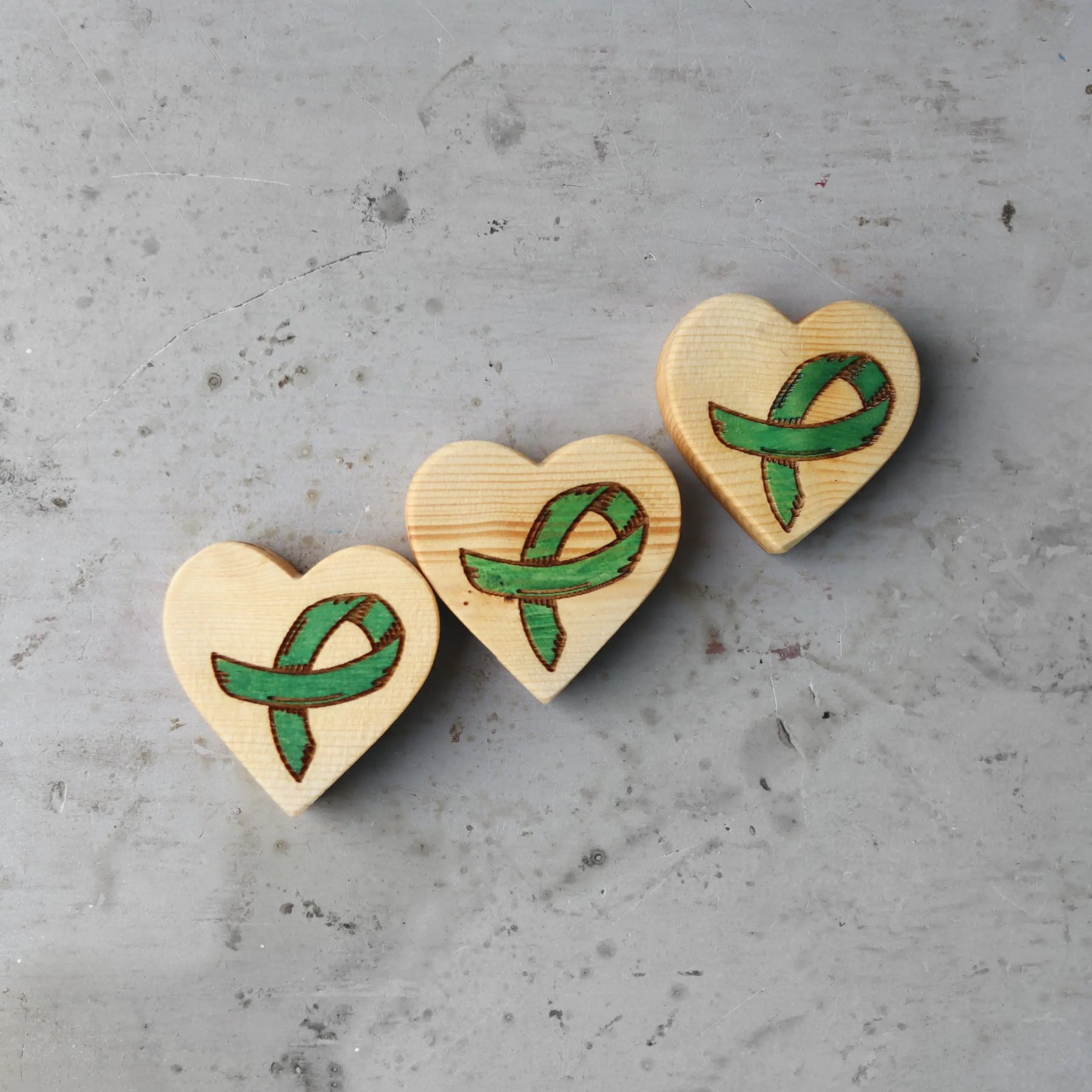Three wooden hearts with green cancer awareness ribbons carved into them on a gray surface.