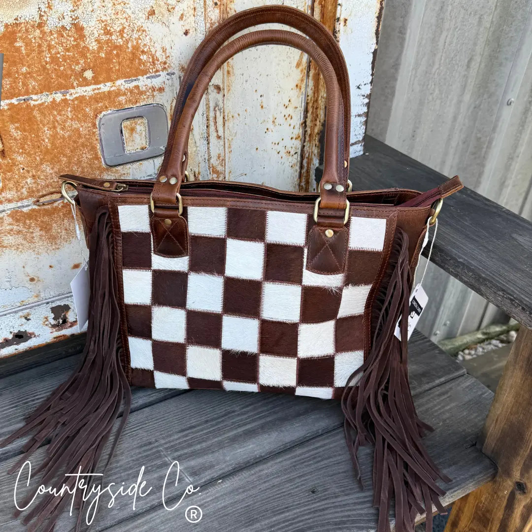 Brown and white cowhide checkerboard purse with fringe and leather handles.