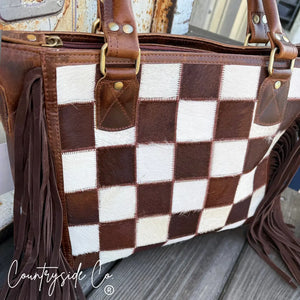 Brown and white checkerboard cowhide purse with leather fringe and handles.
