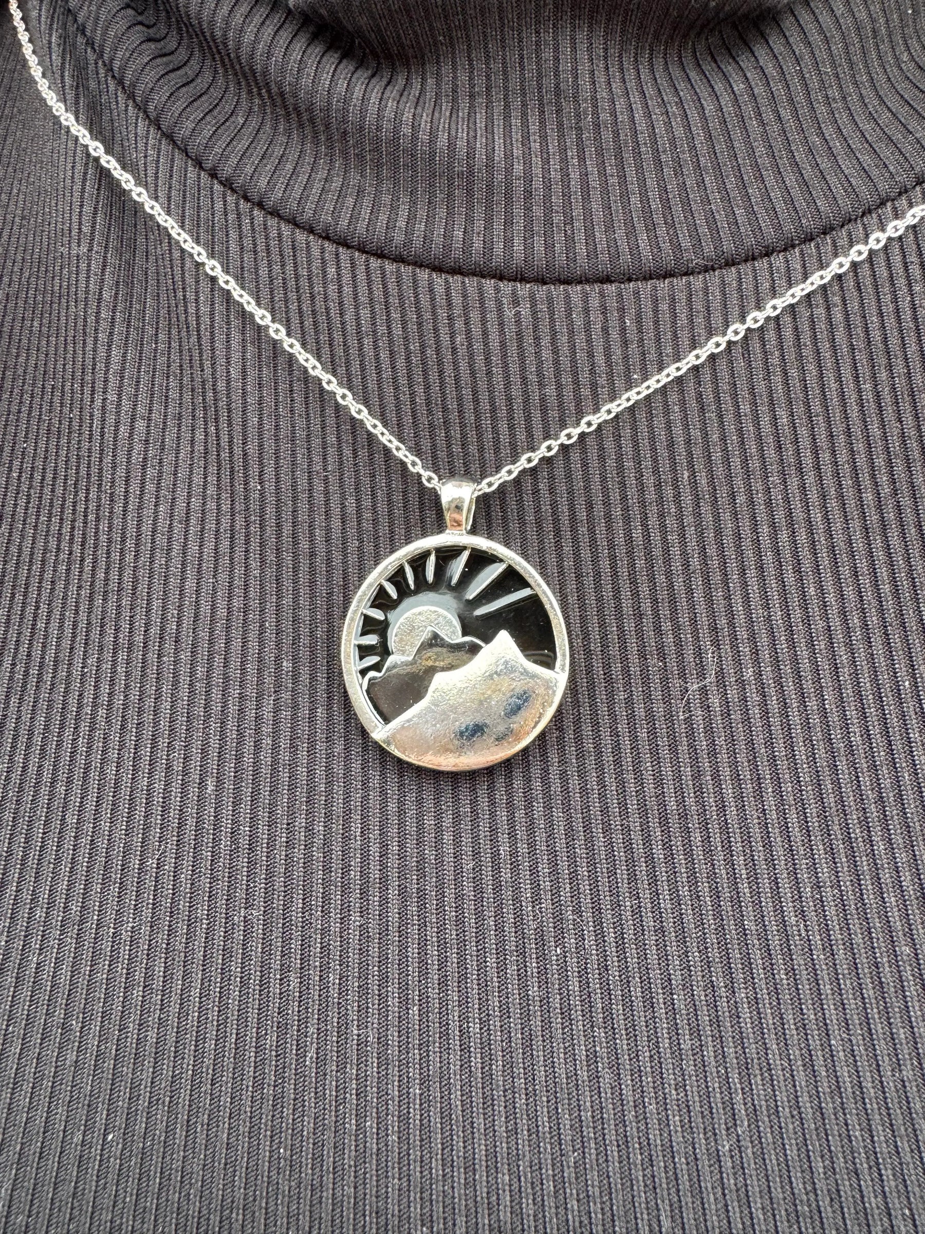 Silver mountain necklace: round pendant with mountain range, sun and navy background on a fine chain against black turtleneck.
