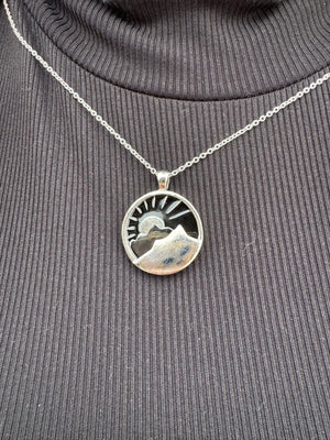 Silver mountain necklace: round pendant with mountain range, sun and navy background on a fine chain against black turtleneck.