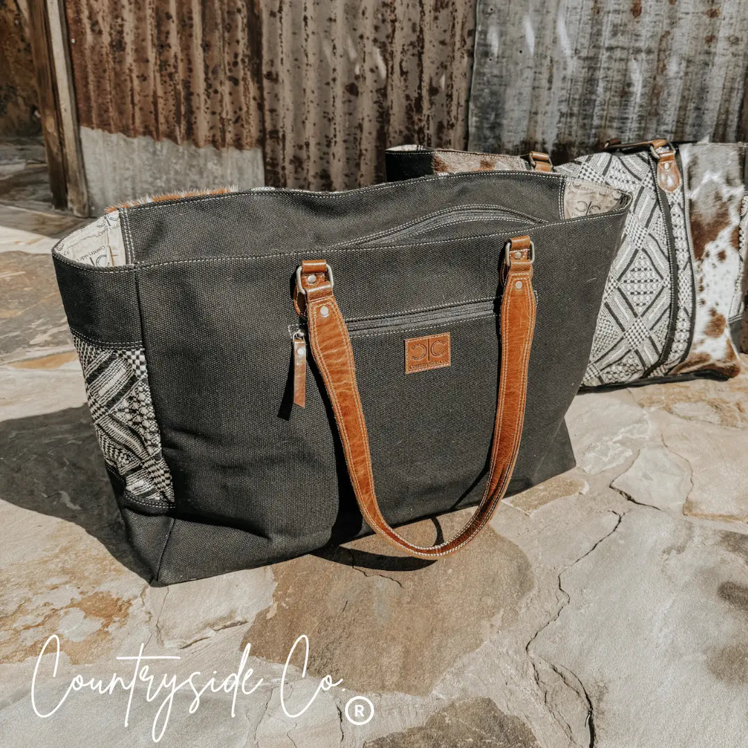 Black canvas weekender bag with cowhide accents, leather handles, and patterned textile sides.