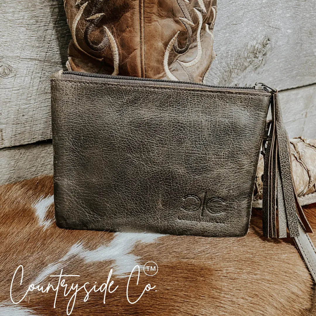 Brown distressed leather wristlet with fringe detail, embossed logo, and stitching next to a cowboy boot.