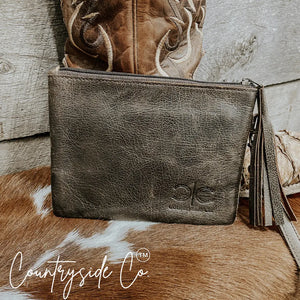 Brown distressed leather wristlet with fringe detail, embossed logo, and stitching next to a cowboy boot.