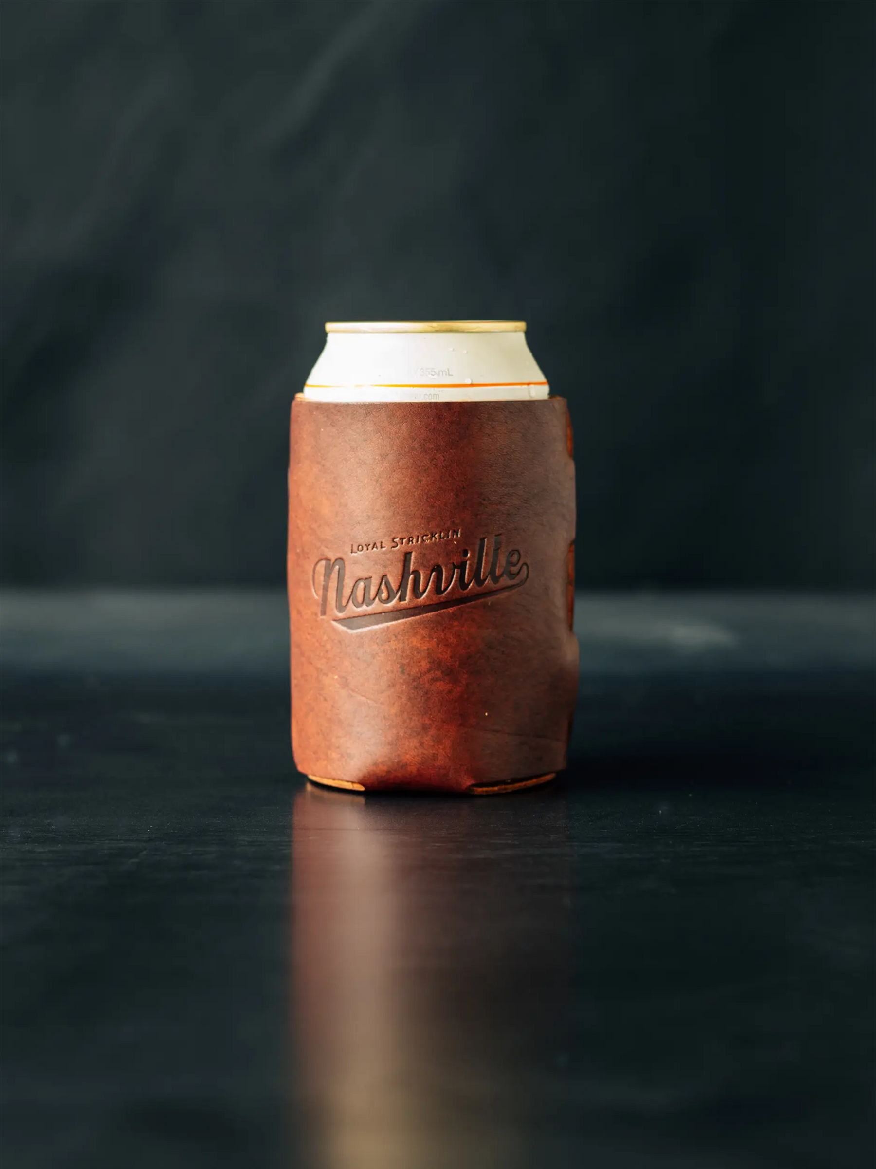 Brown leather coozie with the word "Nashville" on a can of beer on a dark surface.