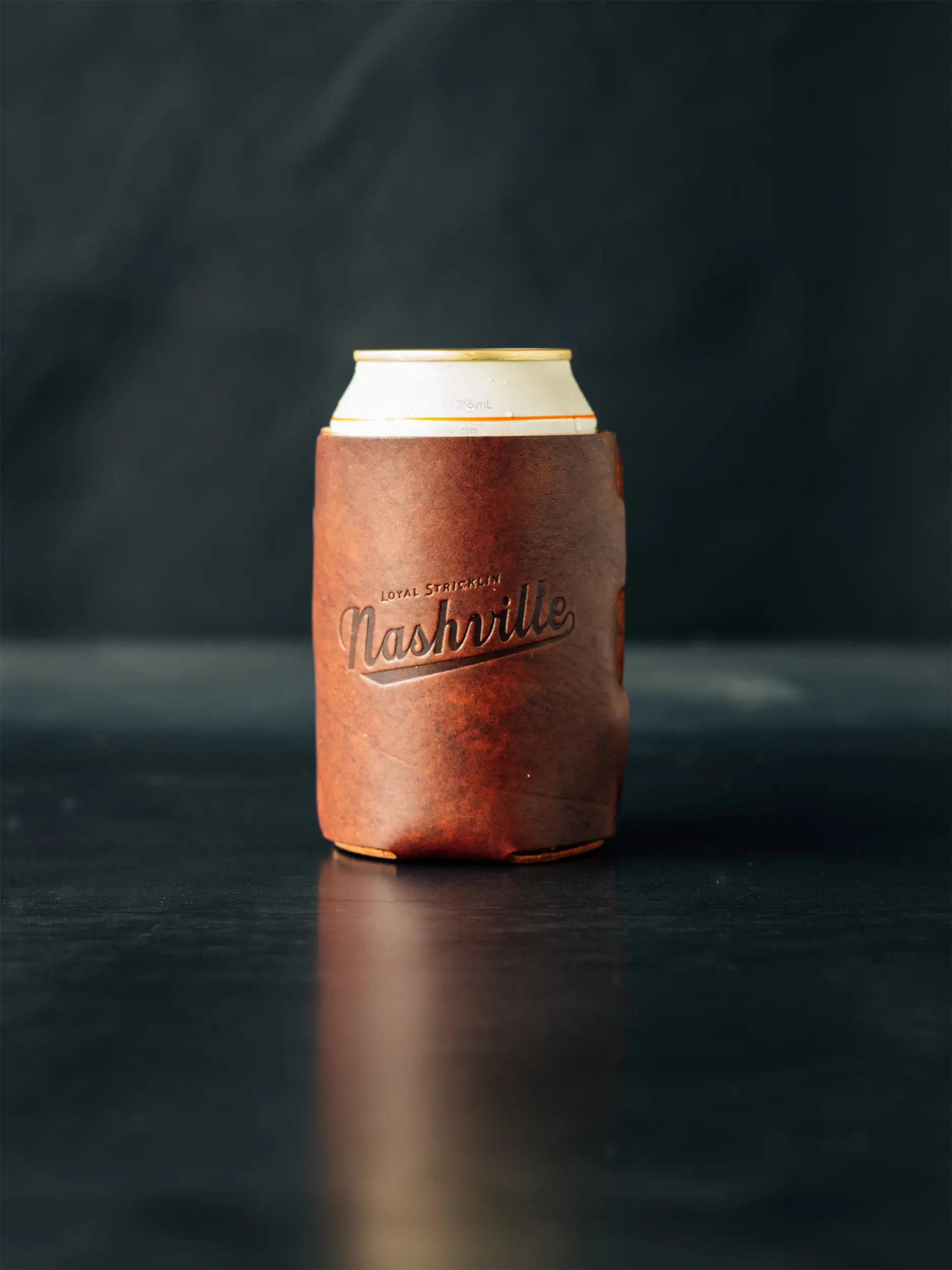 Brown leather coozie with the word "Nashville" on a can of beer on a dark surface.