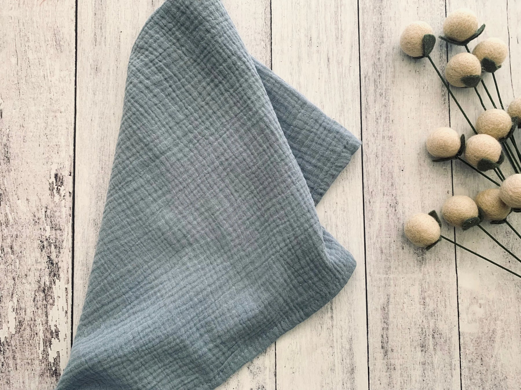 Blue cotton crinkle cloth napkins folded on a white wooden surface with decorative balls on stems.