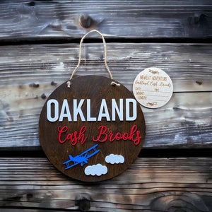 Round wooden baby name announcement sign with airplane and clouds.