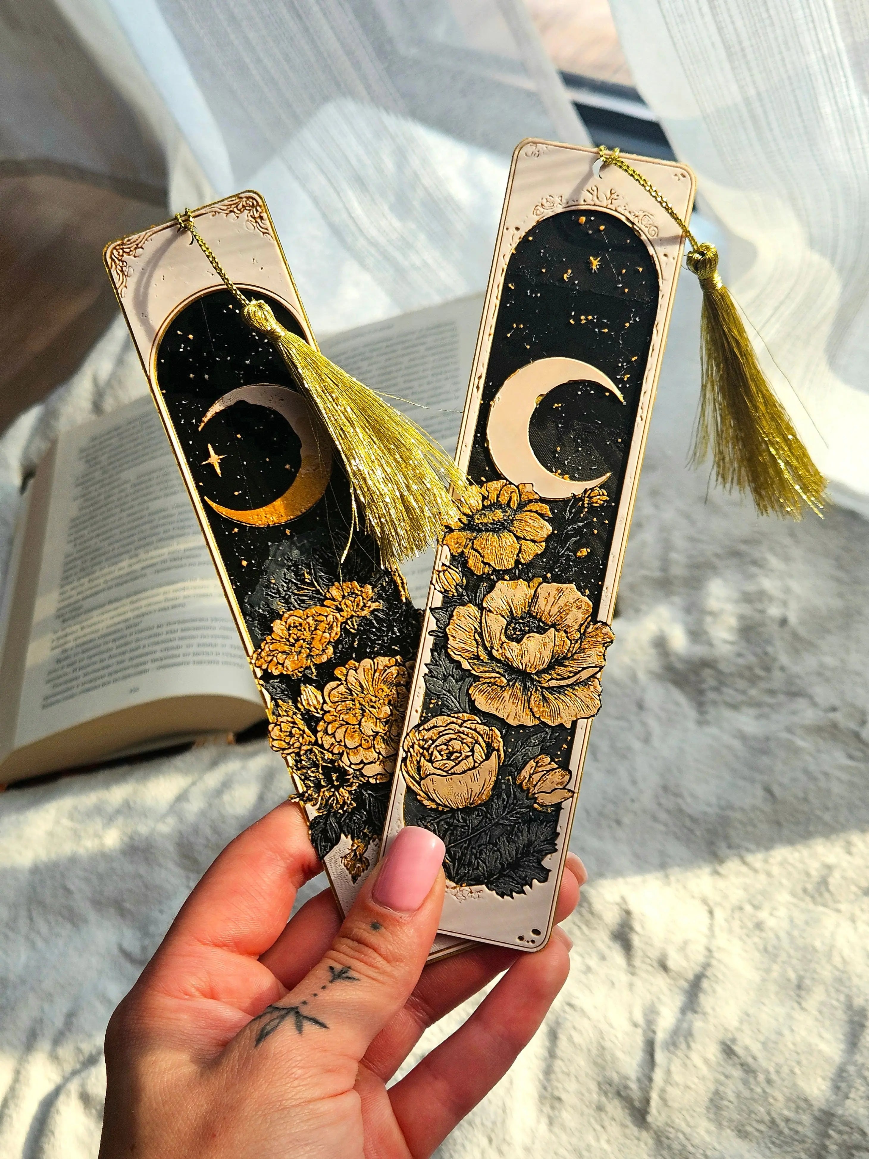 Two celestial bookmarks with crescent moon, stars, floral design, and gold tassels held in hand. Book in background.