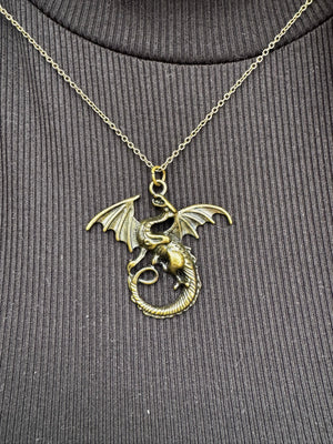 Bronze dragon necklace: a coiled dragon pendant with wings on a gold chain against a dark ribbed knit fabric background.