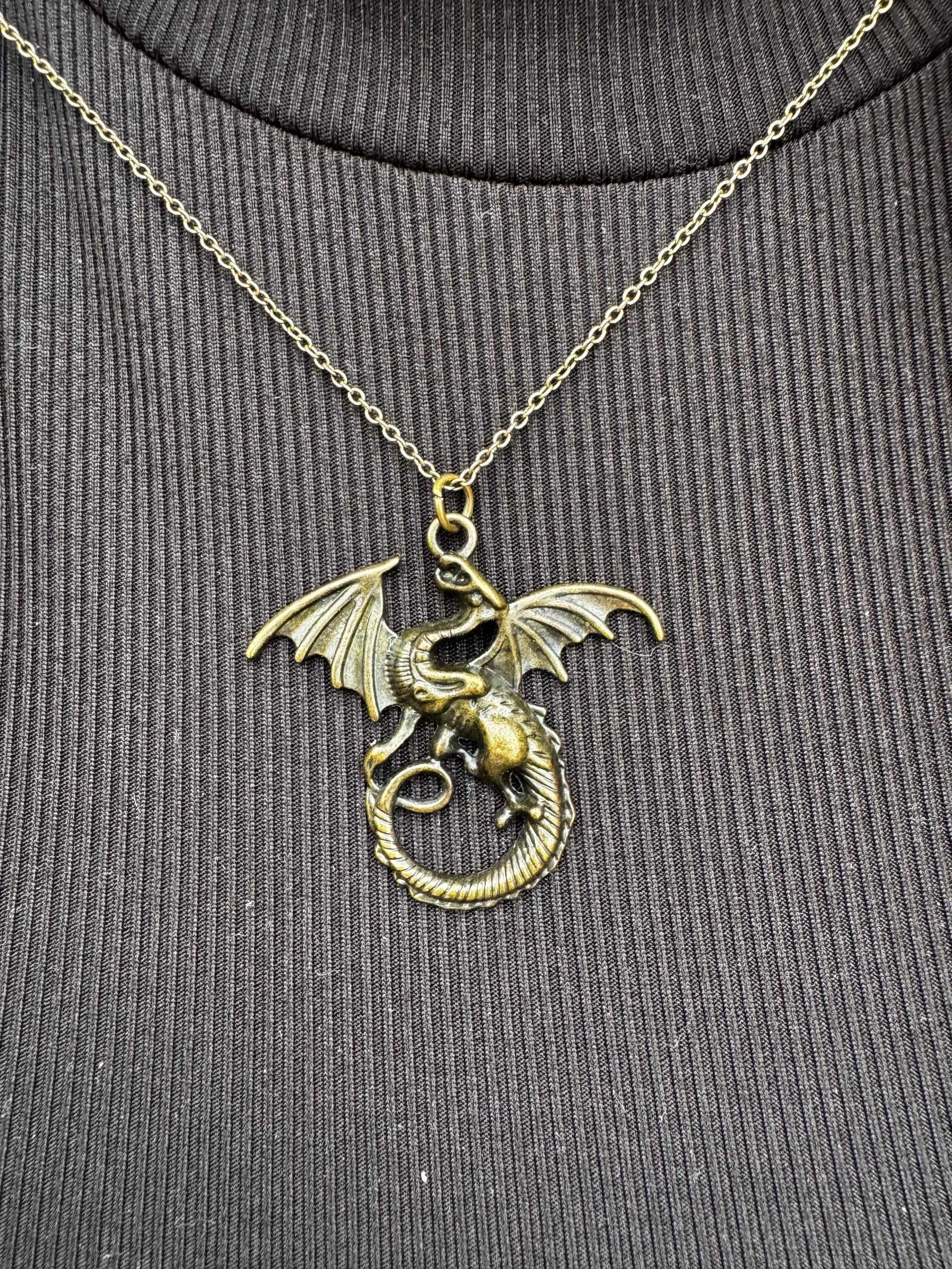 Bronze dragon necklace: a coiled dragon pendant with wings on a gold chain against a dark ribbed knit fabric background.