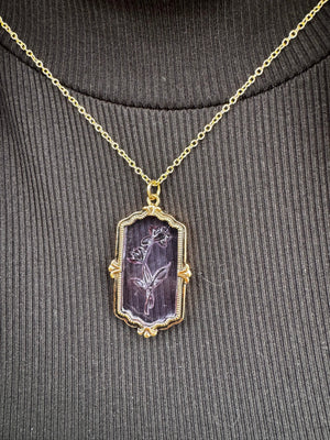 Flower necklace: gold chain, framed purple glass pendant with carved flower on a ribbed dark shirt.