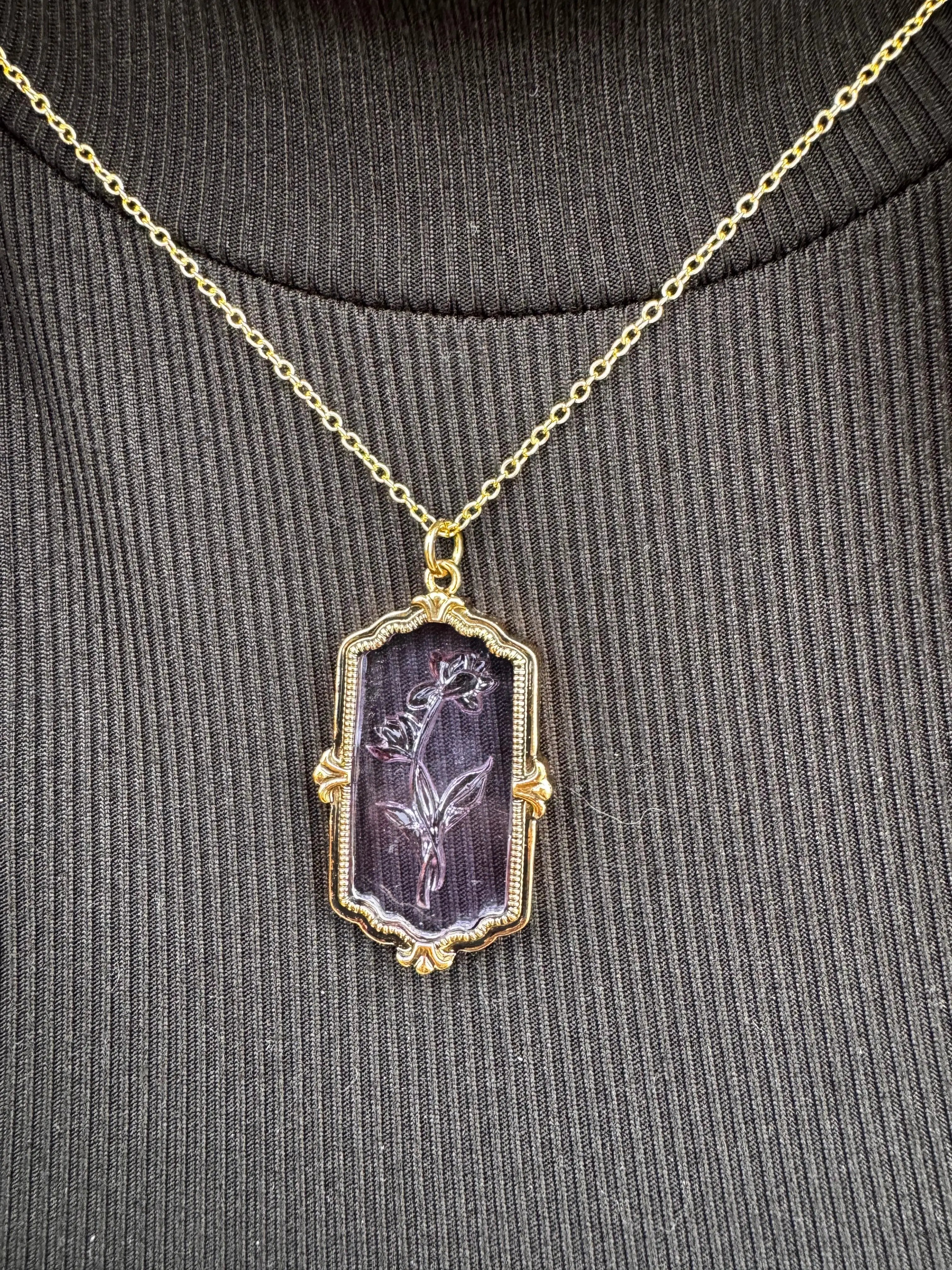 Flower necklace: gold chain, framed purple glass pendant with carved flower on a ribbed dark shirt.