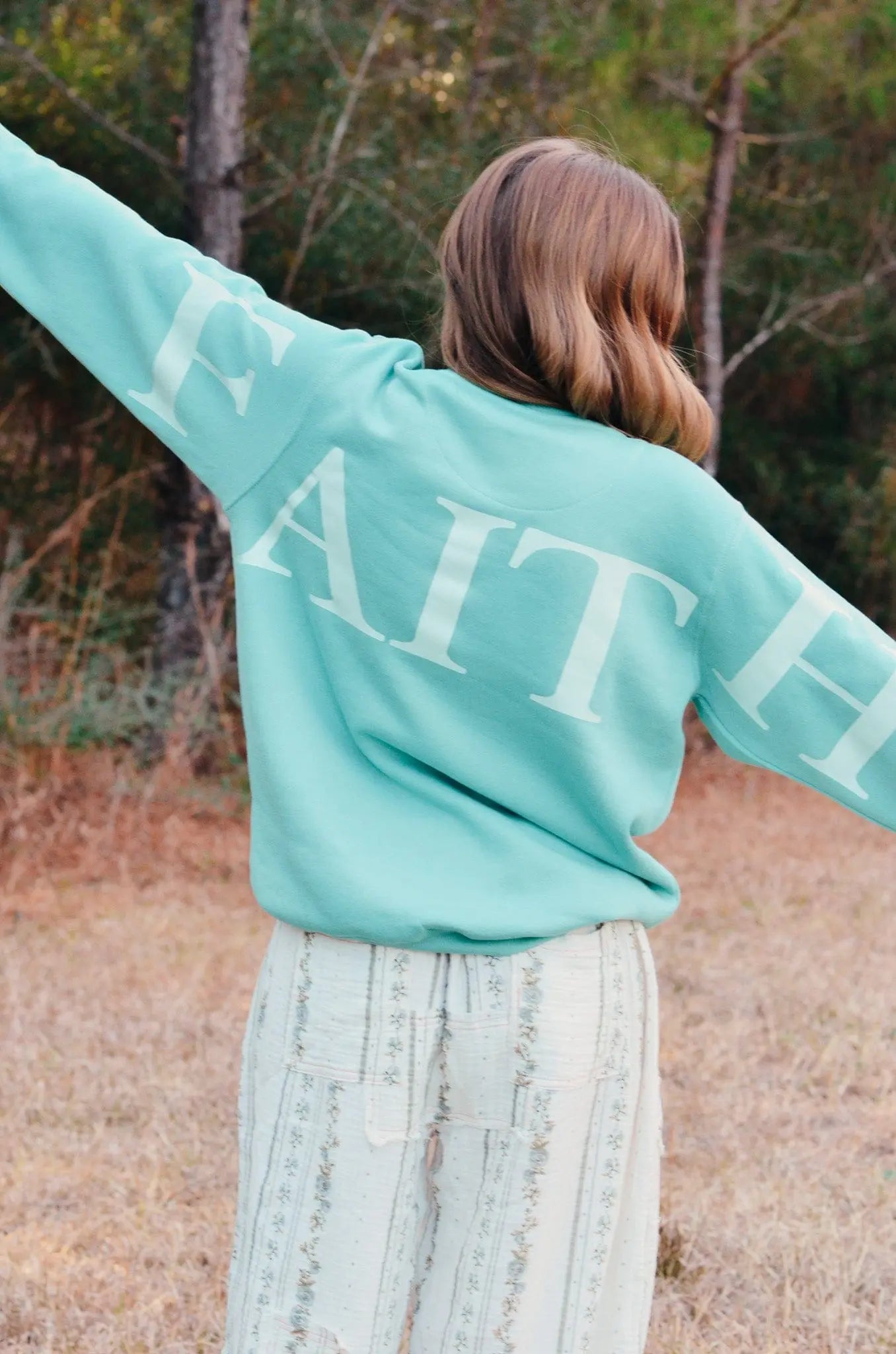 Teal sweatshirt with \"FAITH\" printed on the back and sleeves, worn outdoors.