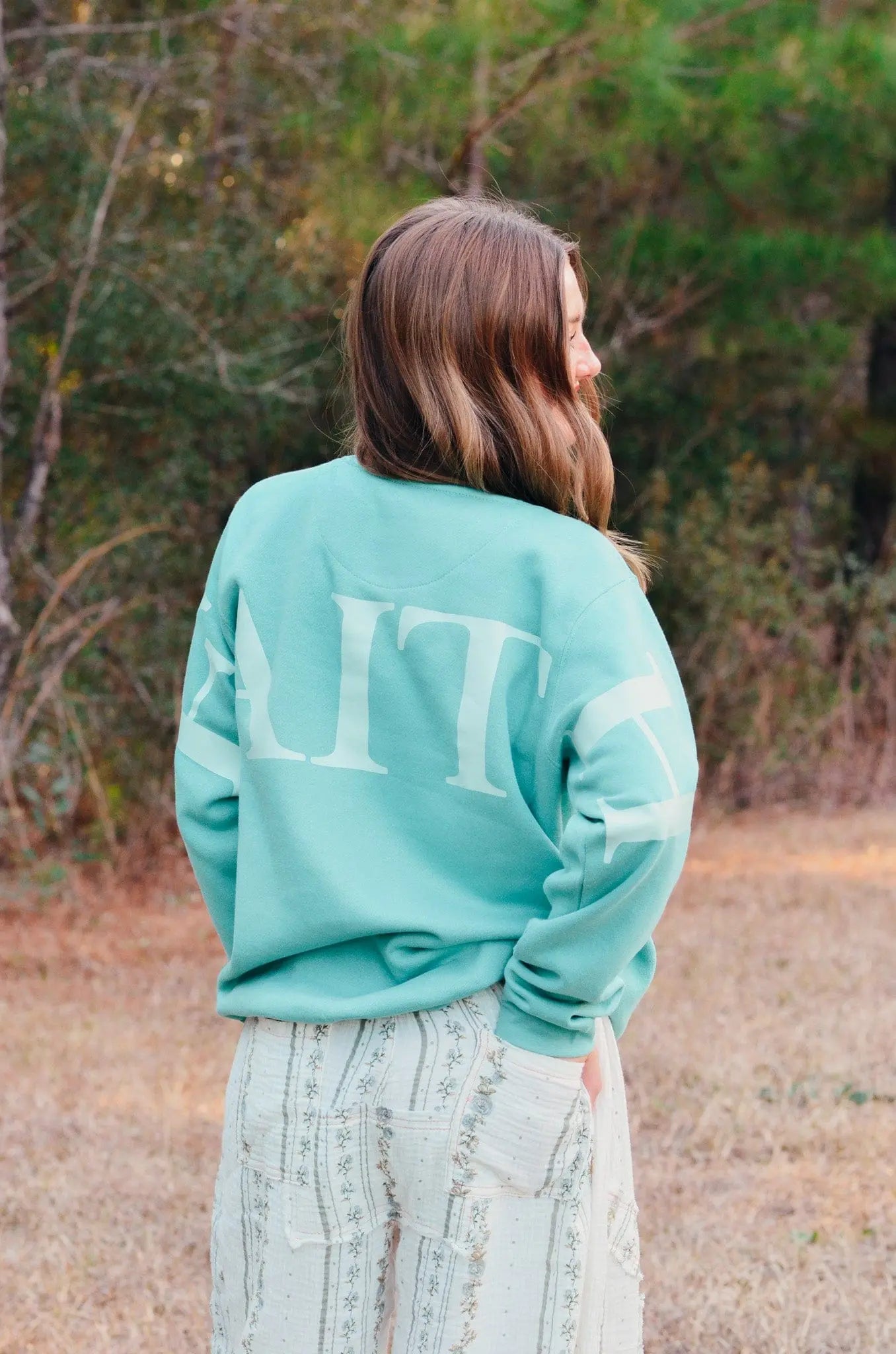 Woman wearing a teal sweatshirt with the word \"FAITH\" printed on the back, posing in an outdoor setting.