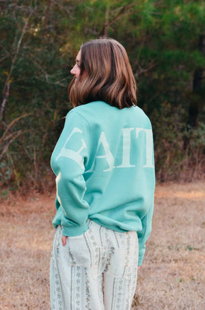Woman with brown hair wearing a teal "Faith" sweatshirt and floral pants, standing in a field with trees in the background.
