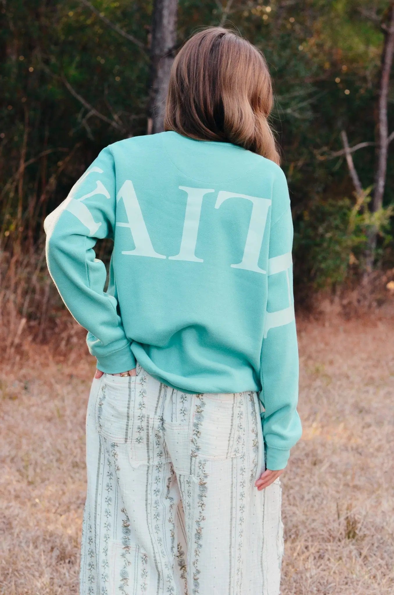 Teal sweatshirt with a white "Faith" back print on a woman standing in a field near trees.